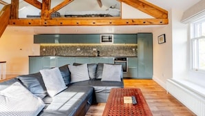 A loft-style room with a sloped ceiling, a built-in kitchen, a sectional sofa, and a wooden coffee table. - Sedbergh Penthouse - KingBed - Tv - Wifi (Sedbergh)
