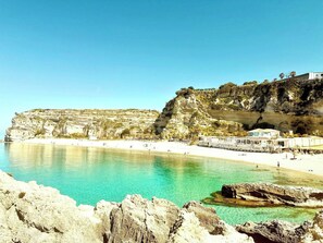 Une zone côtière caractérisée par une plage de sable fin, des falaises rocheuses et un bâtiment à la façade blanche.