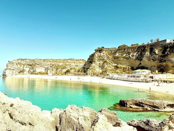 Une zone cÎtiÚre caractérisée par une plage de sable fin, des falaises rocheuses et un bùtiment à la façade blanche.