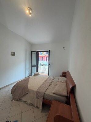 A bedroom with a bed, a wooden bench, a small table, a window with a view of a building, and a ceiling light.