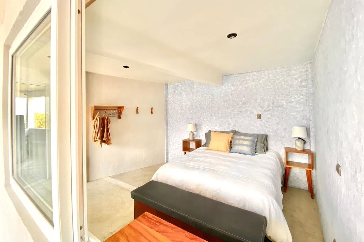 Un dormitorio con un ventanal grande, una cama con una banca, dos mesitas de noche con lámparas y un perchero en la pared.