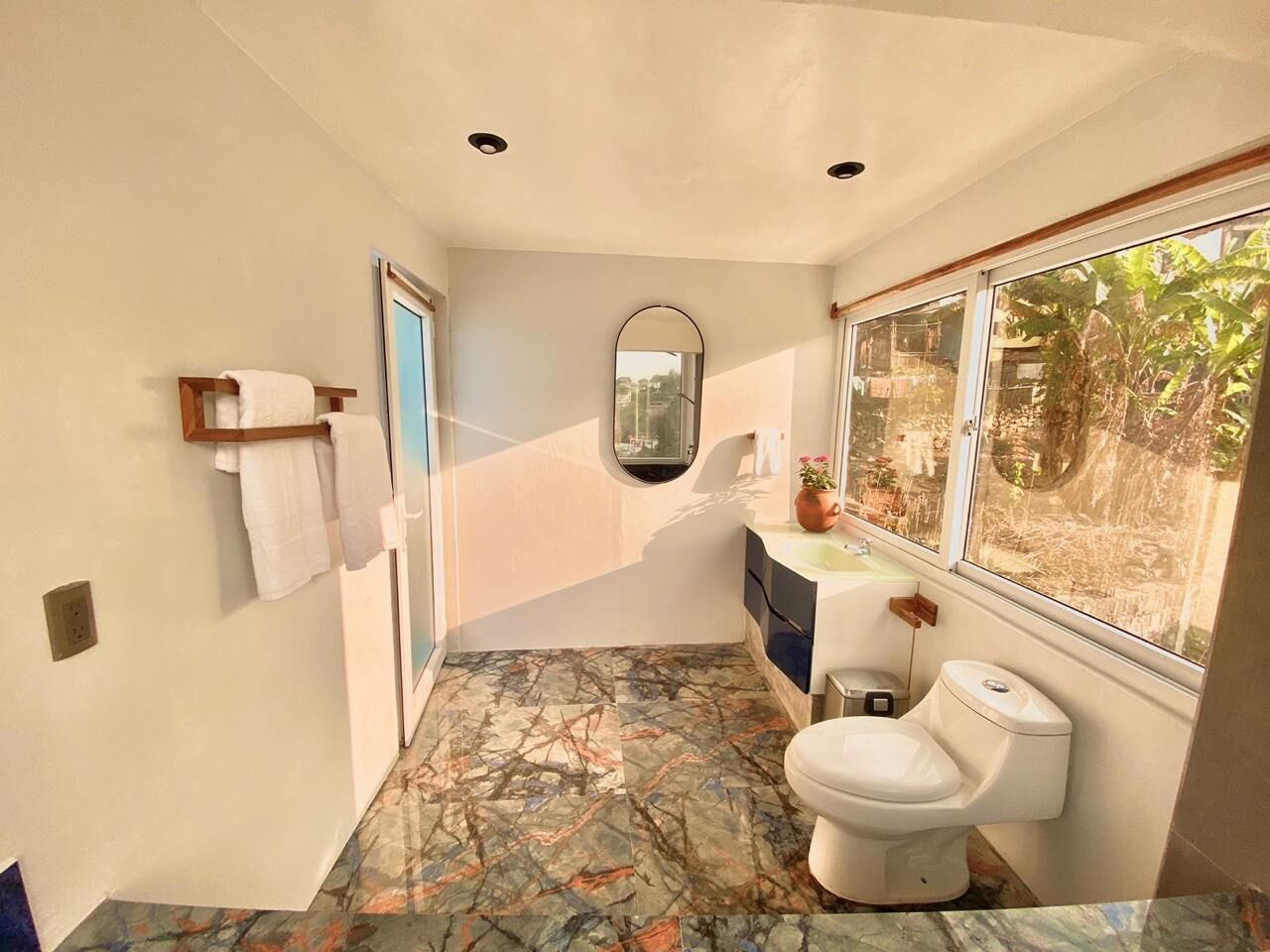A bathroom with a marble floor, a toilet, a sink, a mirror, and a window with a view of greenery.