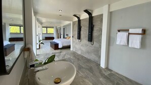 Un baño moderno con un lavamanos grande, una bañera y una zona de ducha. Hay un espejo, un perchero para toallas y un banco. La habitación tiene un ventanal con vistas al exterior.