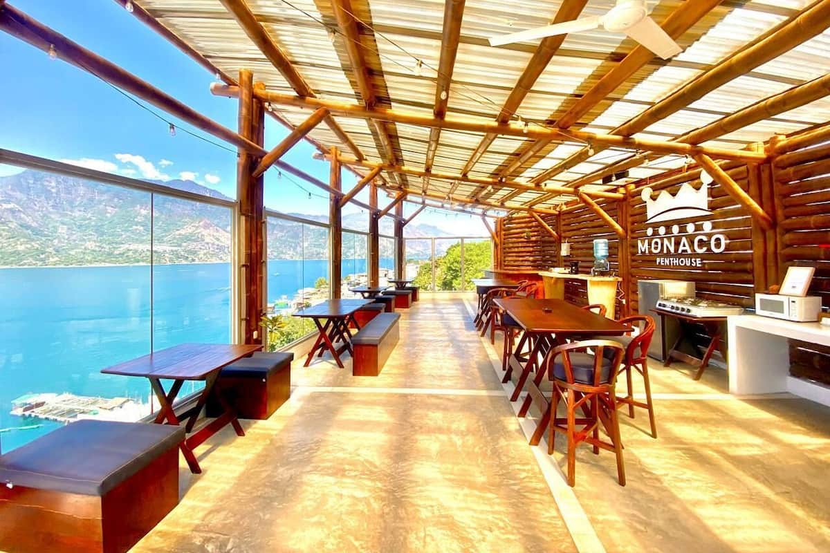 Un restaurante con vistas al mar y a las montañas, con mesas y sillas de madera, una barra con un letrero que dice "Monaco Penthouse" y un amplio ventanal con vista al océano.