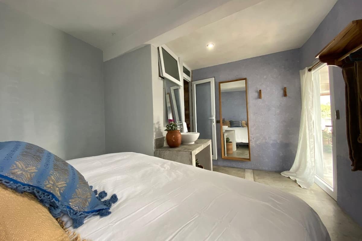 Un dormitorio con una cama, una mesita de noche con un cuenco y una planta, un espejo y una ventana con cortinas.