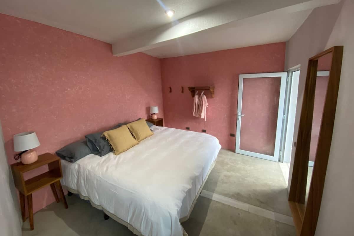 Un dormitorio con una cama grande, una mesita de noche con lámpara, un armario y una puerta que da a otra habitación.