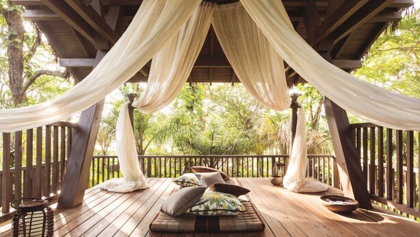A wooden deck with a hammock and cushions, surrounded by trees.