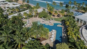 An aerial view of a resort with a central pool, palm trees, and a beach.