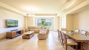 A spacious living room with a large flat-screen TV mounted on a textured wall, a beige sofa set, a wooden coffee table, and a dining area with a glass-top table and wooden chairs.