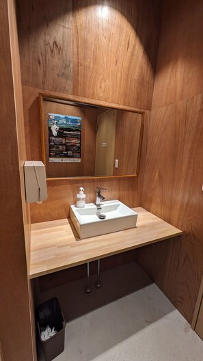 A small, wooden-framed bathroom with a white sink, a mirror, and a poster on the wall. - Aso Sakura Ark - Hostel (Aso)