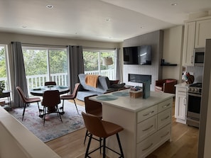 A modern kitchen with a dining area, a flat-screen TV mounted above the counter, and a large window with curtains.
