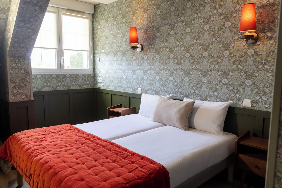 Un lit double avec une couverture rouge, deux tables de chevet, une fenêtre et un papier peint à motifs.