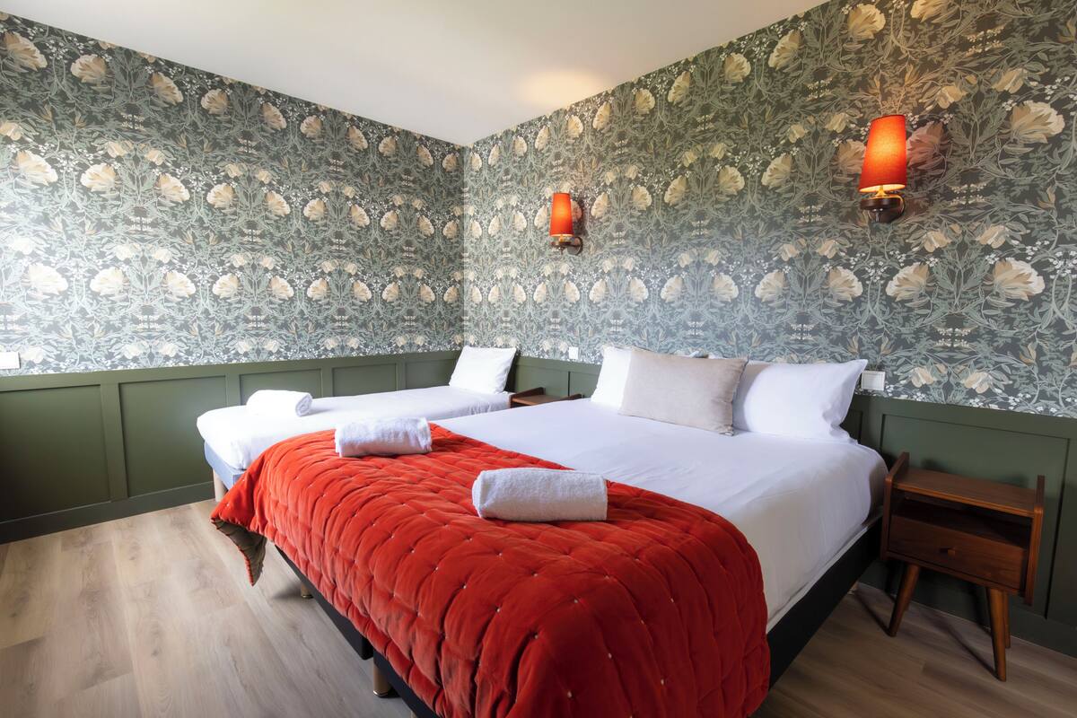 Un lit double avec une couverture rouge, deux tables de chevet et un papier peint à motifs floraux.