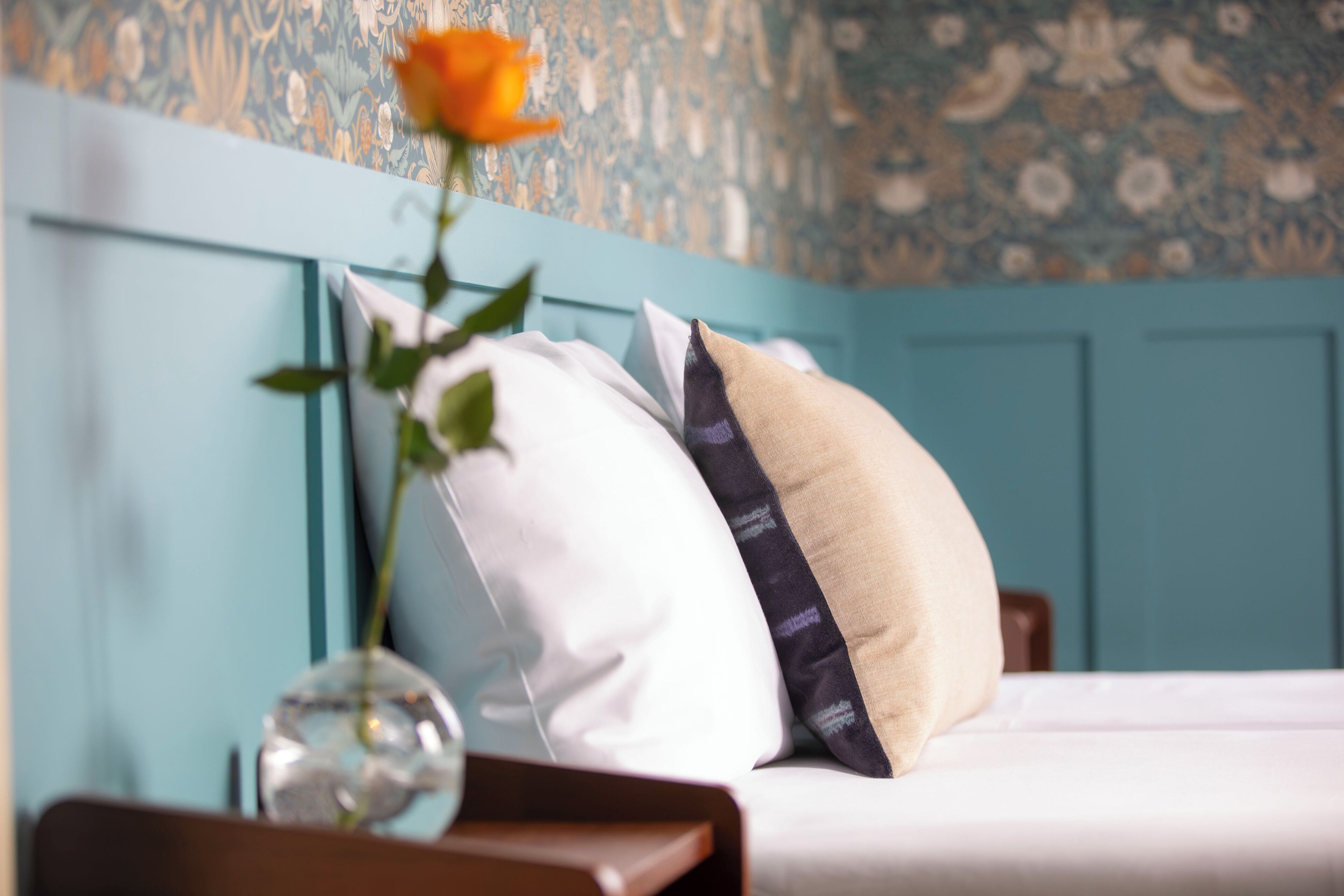 Uma cama com almofadas brancas e bege, um vaso com uma flor e uma parede azul com papel de parede floral.