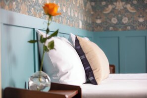A bed with white and beige pillows, a vase with a flower, and a blue wall with floral wallpaper. - Relais Touraine Sologne (Noyers-sur-Cher)
