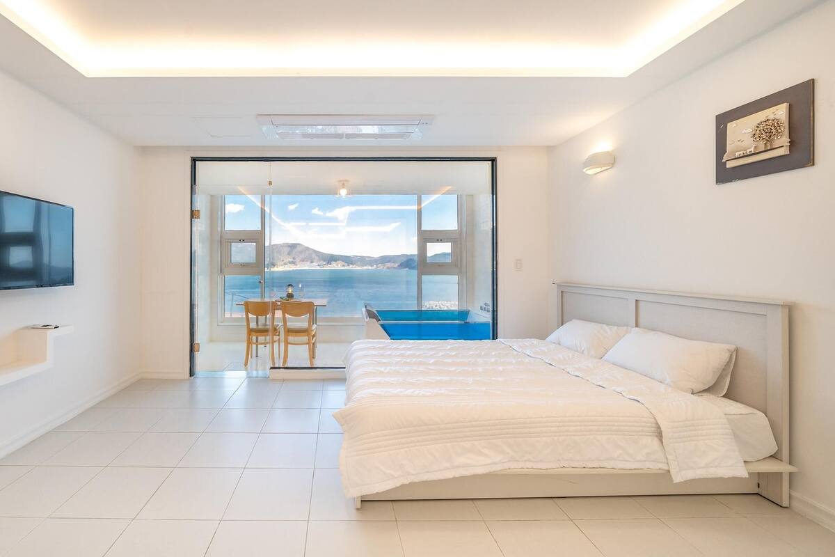 A modern bedroom with a bed, a TV, a view of the sea, and a balcony with a table and chairs.