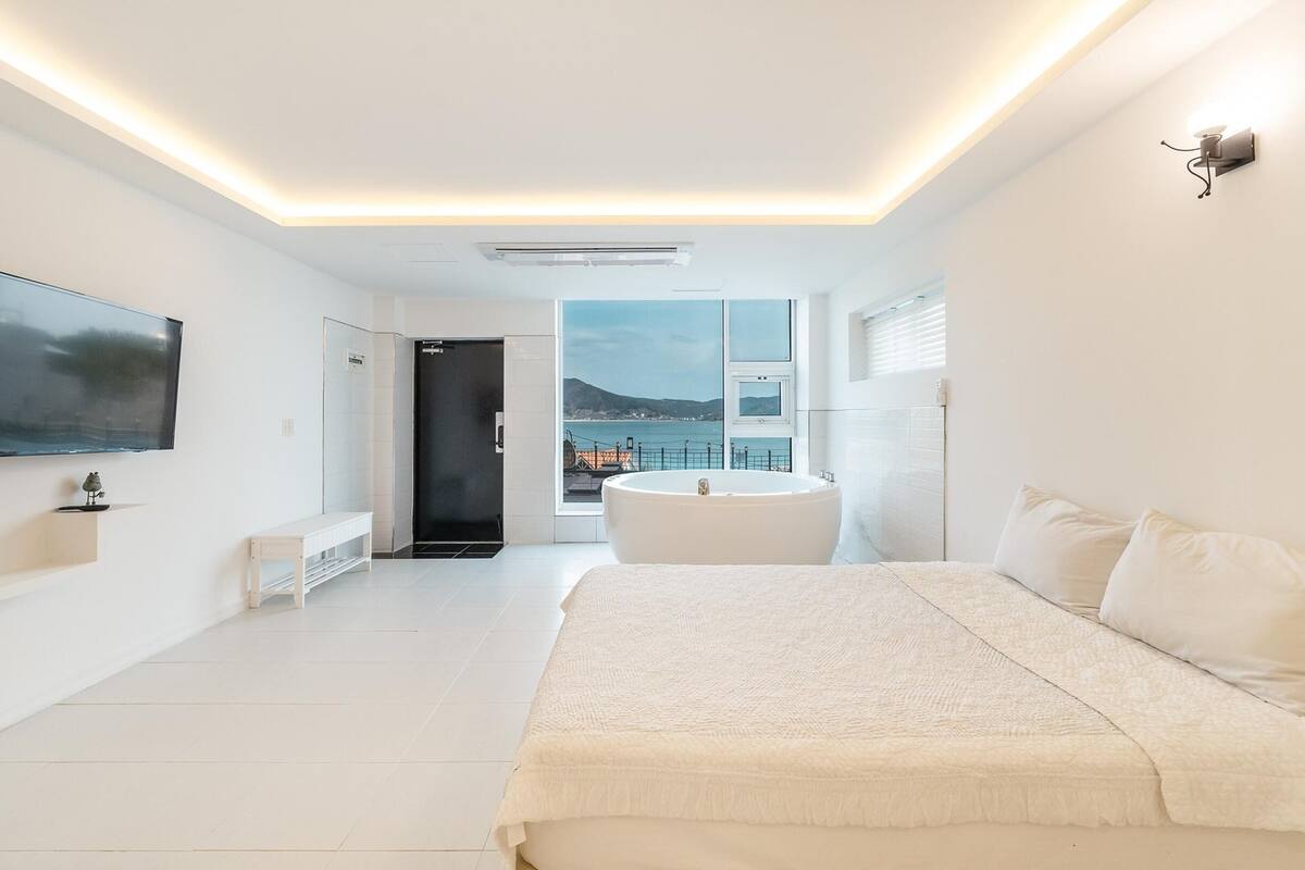 A modern bedroom with a large bed, a flat-screen TV, a bathtub, and a view of the sea.
