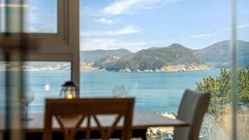 A dining area with a table and chairs, overlooking a coastal landscape with hills and a body of water.