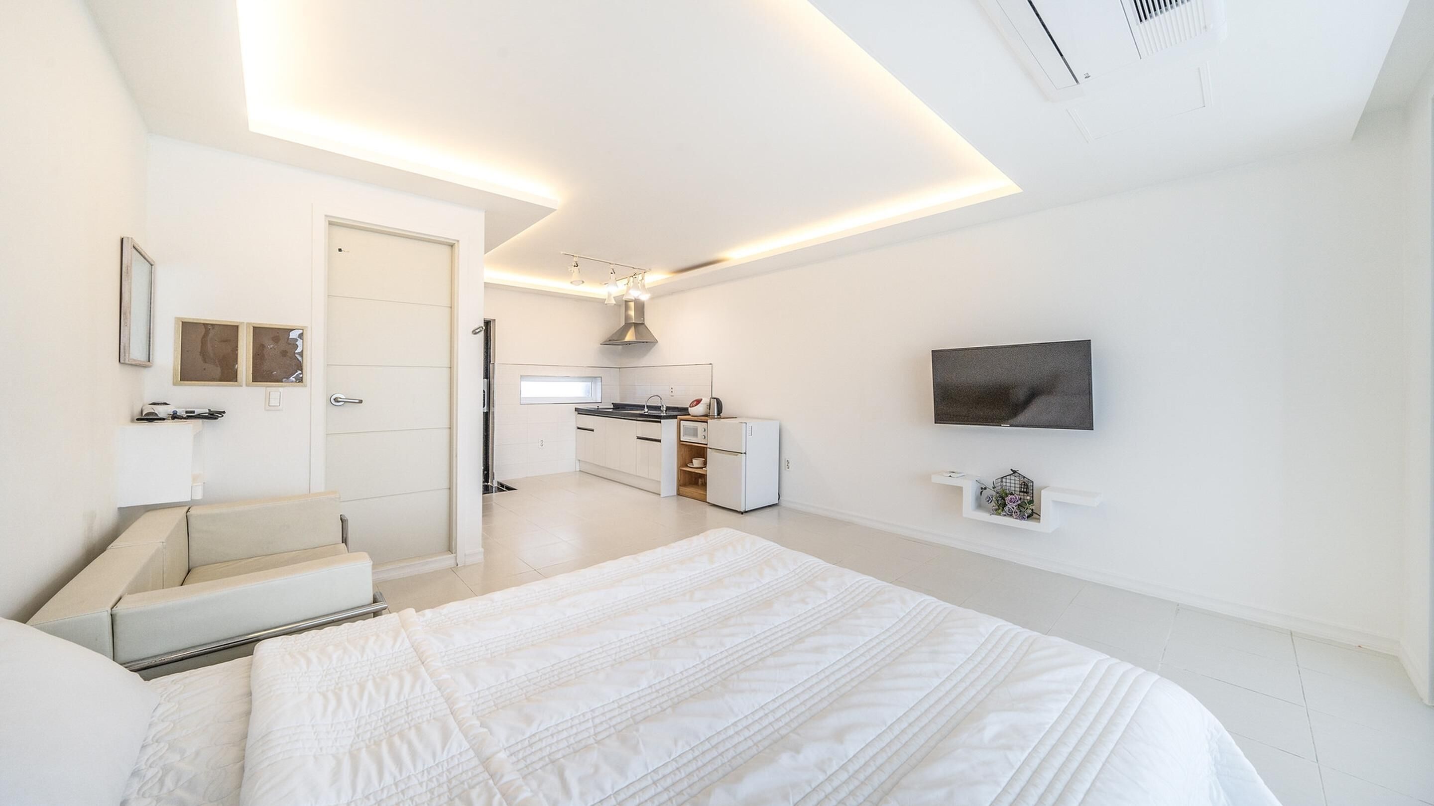 A modern bedroom with a large bed, a TV mounted on the wall, and a kitchenette in the background.