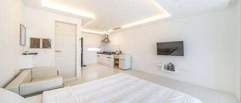 A modern bedroom with a large bed, a TV mounted on the wall, and a kitchenette in the background.