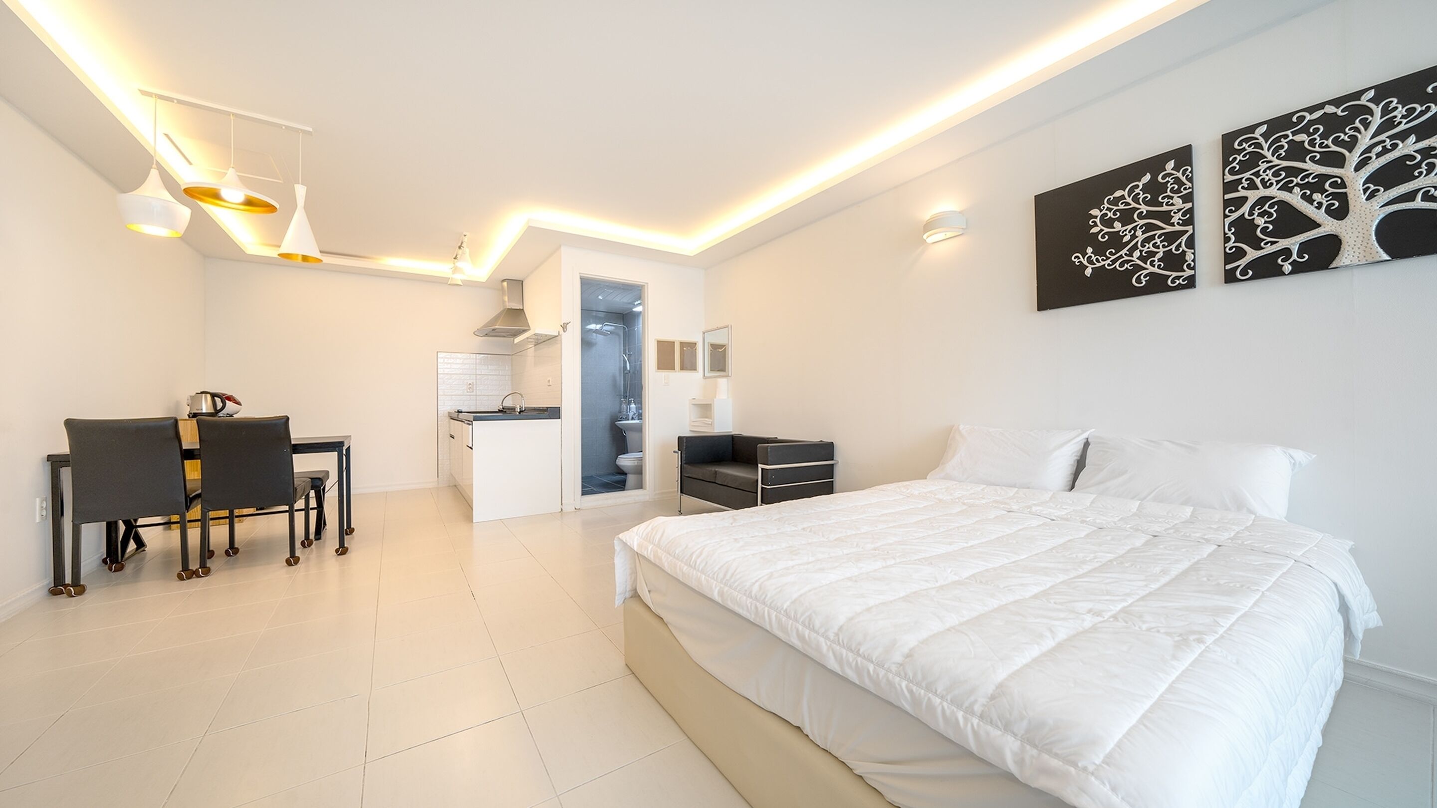 A modern bedroom with a large bed, a desk with chairs, a kitchenette, and a bathroom.