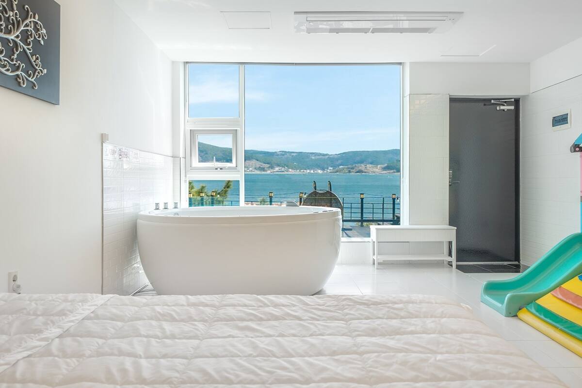 A bedroom with a large bed, a bathtub, a small table, a colorful play structure, and a view of the sea.