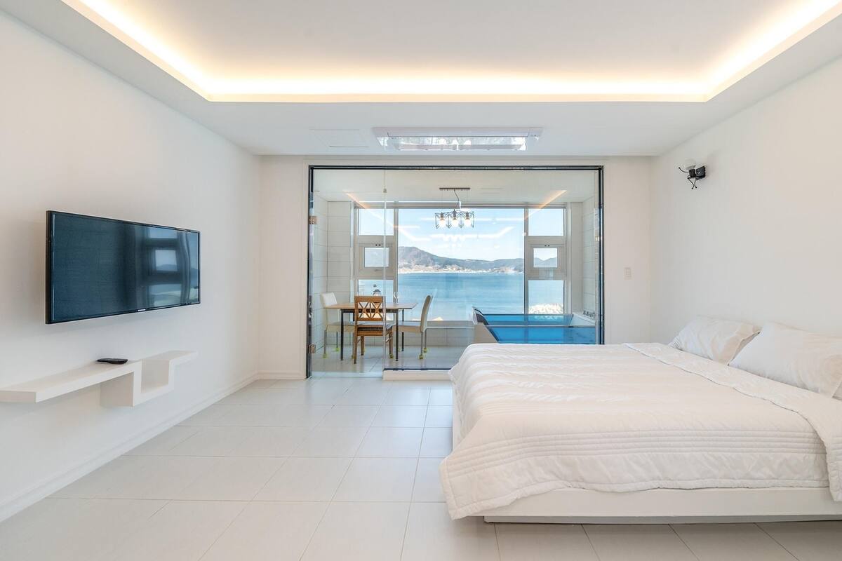 A modern bedroom with a large bed, a flat-screen TV, a dining area with a view of the sea, and a glass door leading to a balcony.