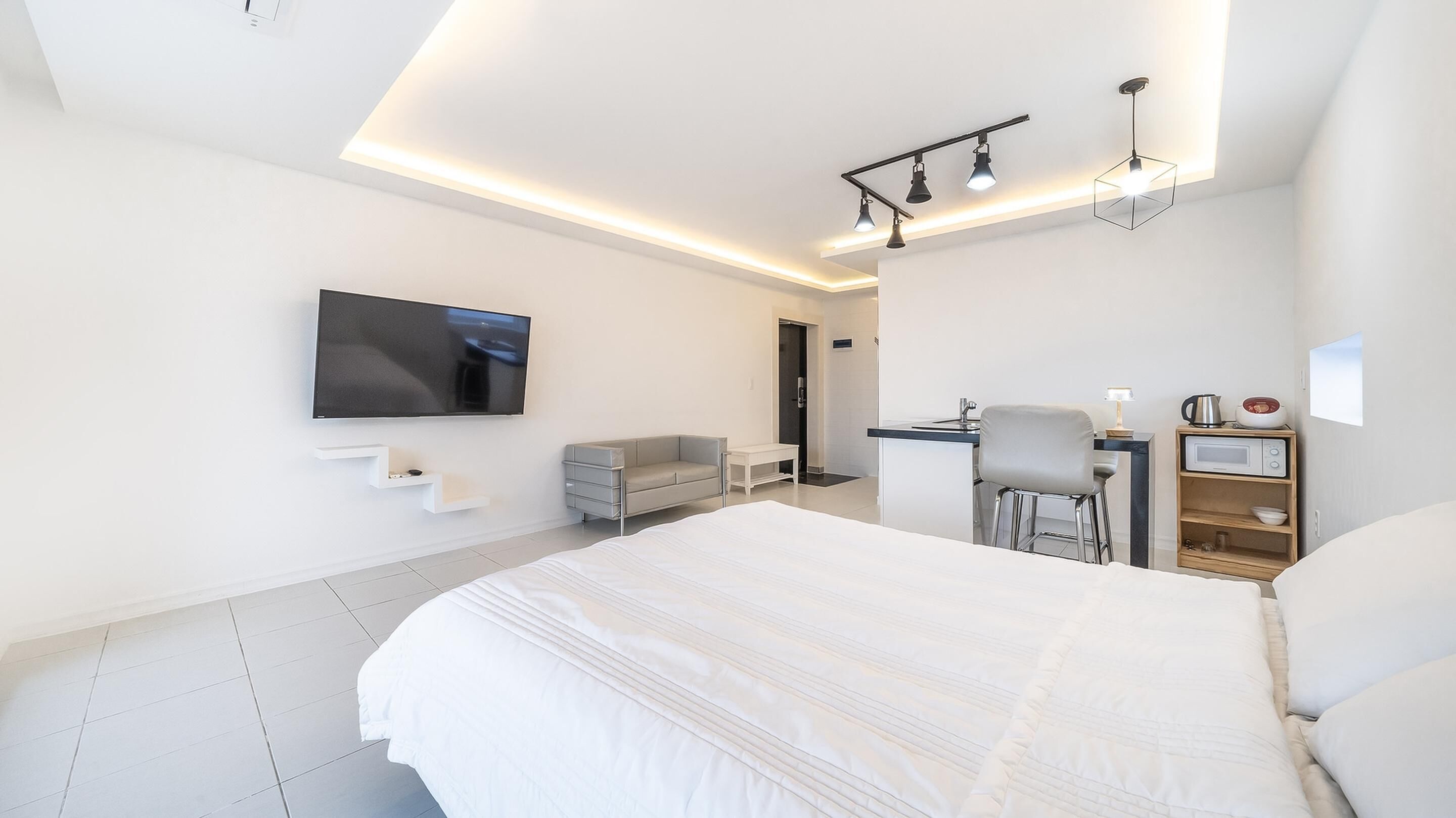 Una habitación de hotel moderna con una cama grande, un televisor de pantalla plana montado en la pared, un escritorio con una silla y una pequeña kitchenette.
