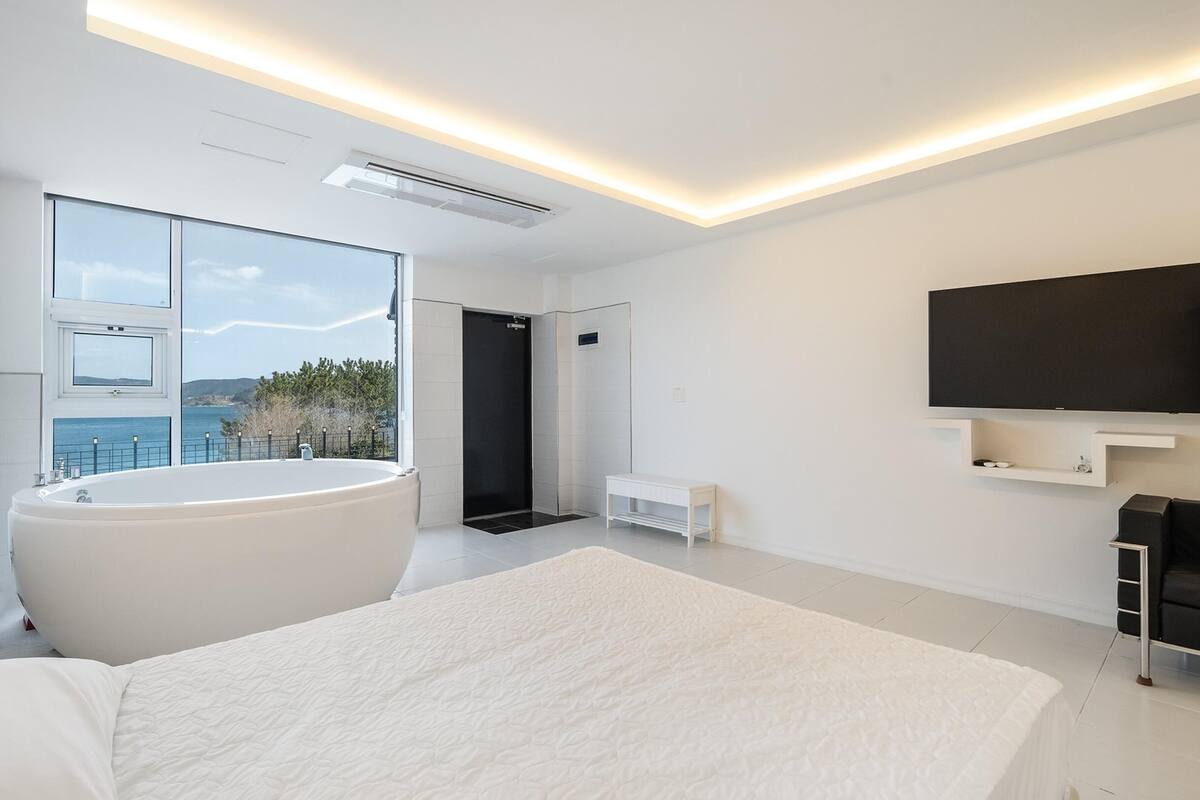 A modern bedroom with a large bed, a bathtub, a TV, and a view of the sea.