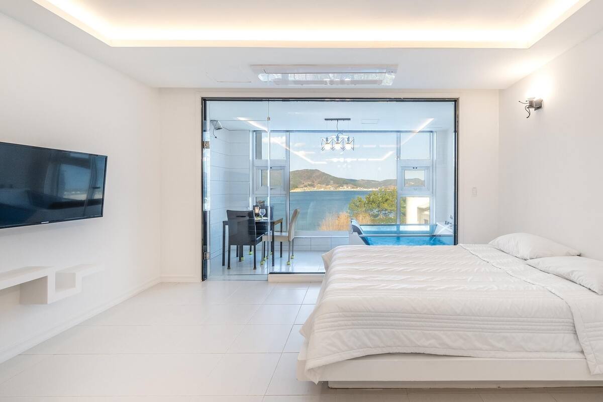 A modern bedroom with a large bed, a TV mounted on the wall, and a view of a pool and mountains through a glass door.