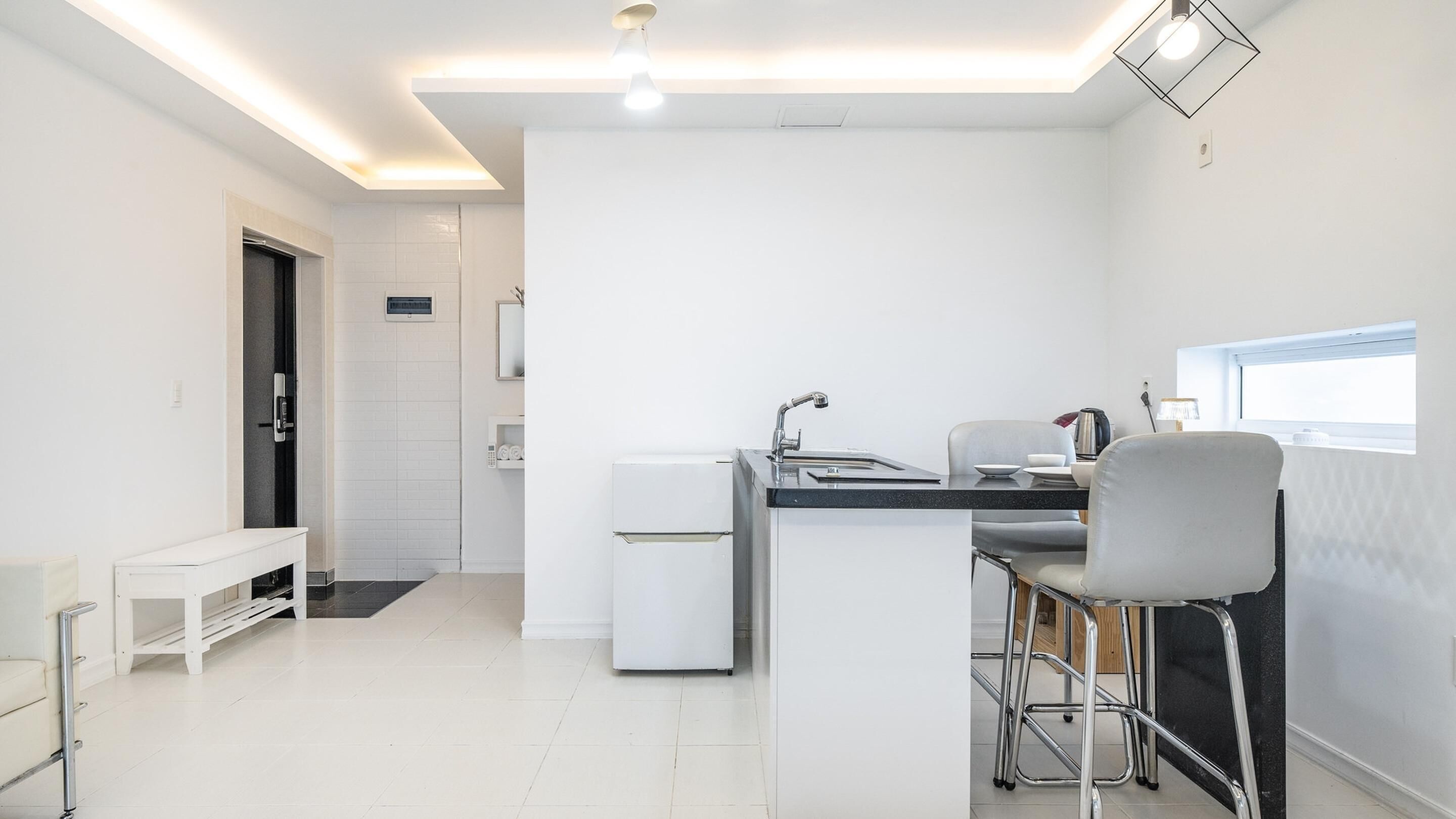 A modern kitchen with a central island, a built-in sink, and a small refrigerator. There is a bench and a window providing natural light.