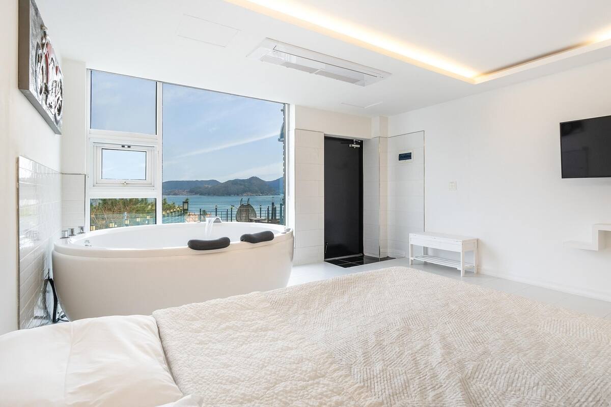 A modern bedroom with a large bed, a bathtub, a TV, and a view of the sea.