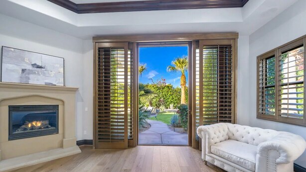 A room with a fireplace, a white sofa, and a view of a garden through open shutters.