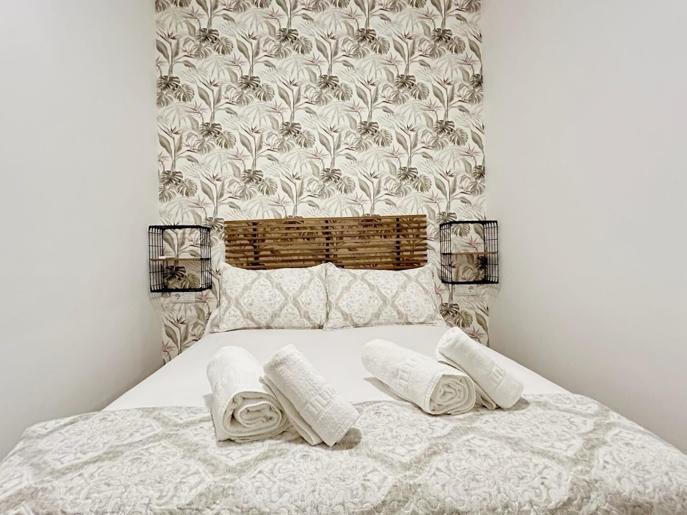 A neatly made bed with rolled white towels, a wooden headboard, and a floral-patterned wall.
