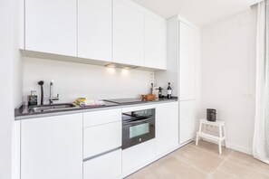 Cozinha moderna com armários brancos, forno embutido, pia e um banquinho branco.