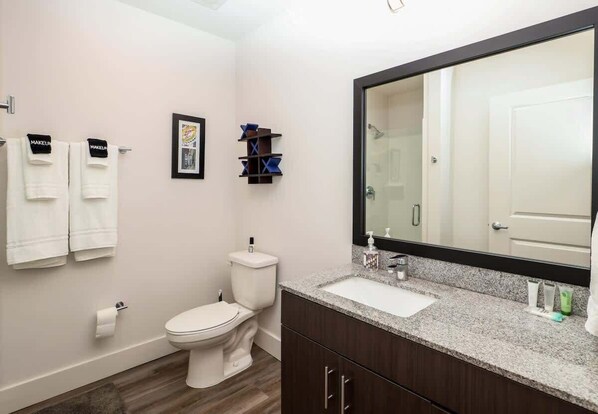 A modern bathroom with a toilet, sink, and large mirror. There are towels hanging on the wall and a framed picture above the toilet.
