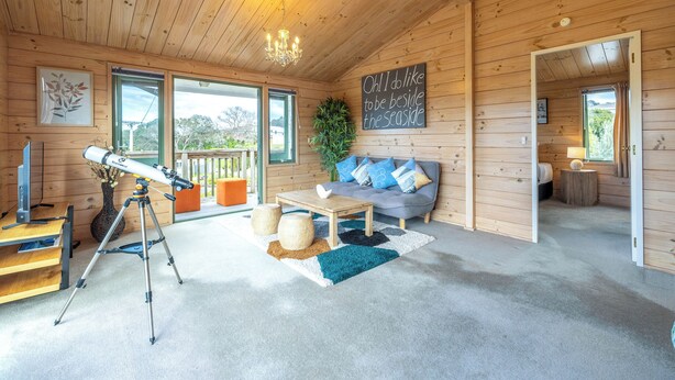 A cozy cabin interior with a telescope, a sofa, and a large window offering a view of the outdoors. -  located in the heart of Oneroa, landscaped garden and 5 minute's walk to beach (Waiheke Island)