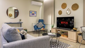 A living room with a grey sofa, a blue armchair, a coffee table, a TV, and a wall-mounted air conditioner. - Bubble Beach 2-BR in Souk Batroun with Parking
 (Batroun)