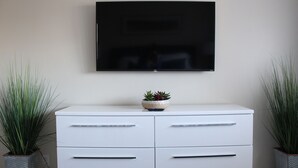 Ein weißes Schränkchen mit Schubladen und ein Flachbildfernseher, der darüber an der Wand angebracht ist. Auf jeder Seite des Schränkchens stehen je ein Blumentopf.