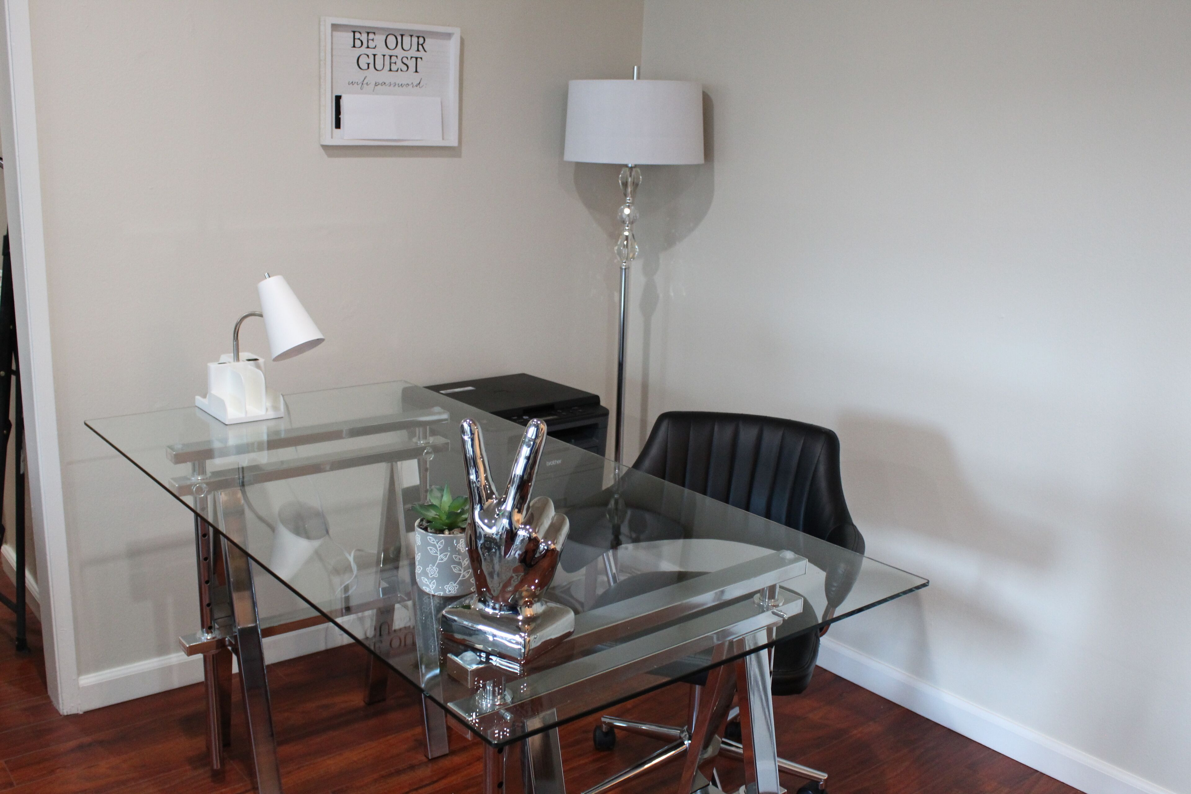 Un espace de bureau moderne avec une table en verre, un fauteuil de bureau noir, une lampe de bureau blanche et un panneau encadré accroché au mur.