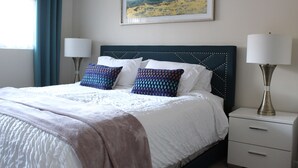 Ein ordentlich bezogenes Bett mit einem gemusterten Kopfteil, ein Nachttisch mit einer Lampe und ein gerahmtes Kunstwerk an der Wand.