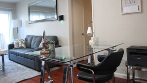 Ein modernes Wohnzimmer mit einem Glastisch, einem schwarzen Ledersofa, einem Schreibtisch mit Glasplatte und einem Stuhl sowie einem großen Spiegel an der Wand.