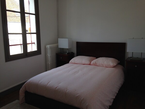 A bedroom with a bed, two bedside tables with lamps, a radiator, and a window with blinds.