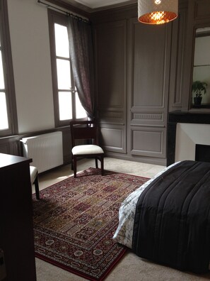 A bedroom with a large bed, a chair, a radiator, a window with curtains, and a hanging light fixture. - Maison du Chatelain - Zoo de Beauval (Saint-Aignan)
