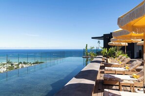 A rooftop infinity pool with a view of the ocean, wooden lounge chairs, and yellow umbrellas. - Legend Hill Residences & Spa (Black River)