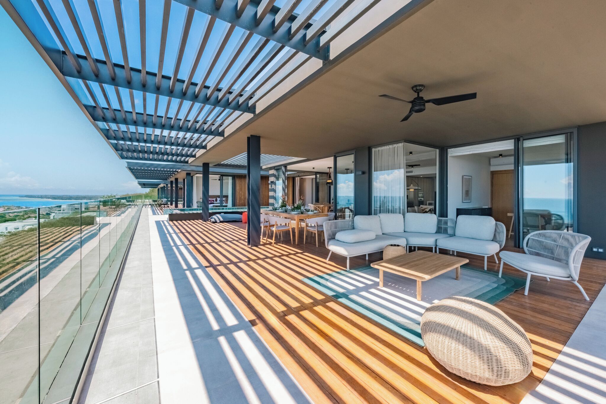 A modern outdoor balcony with a wooden floor, white sofas, a dining table, and a ceiling fan.