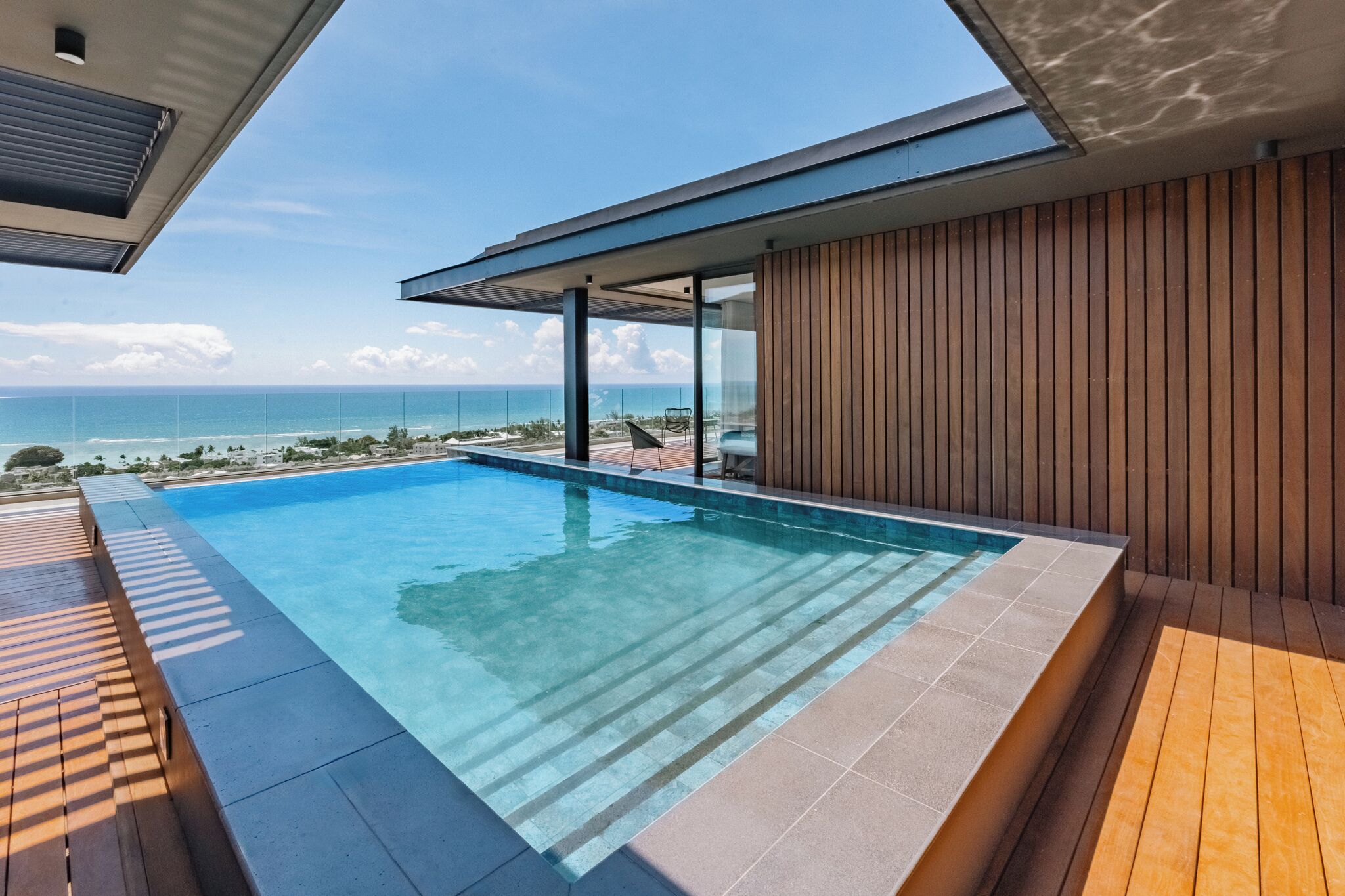 Piscina infinita moderna com vista clara do oceano, deck de madeira e área envidraçada com vista para a praia.