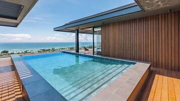 Piscina infinita moderna com vista clara do oceano, deck de madeira e área envidraçada com vista para a praia.