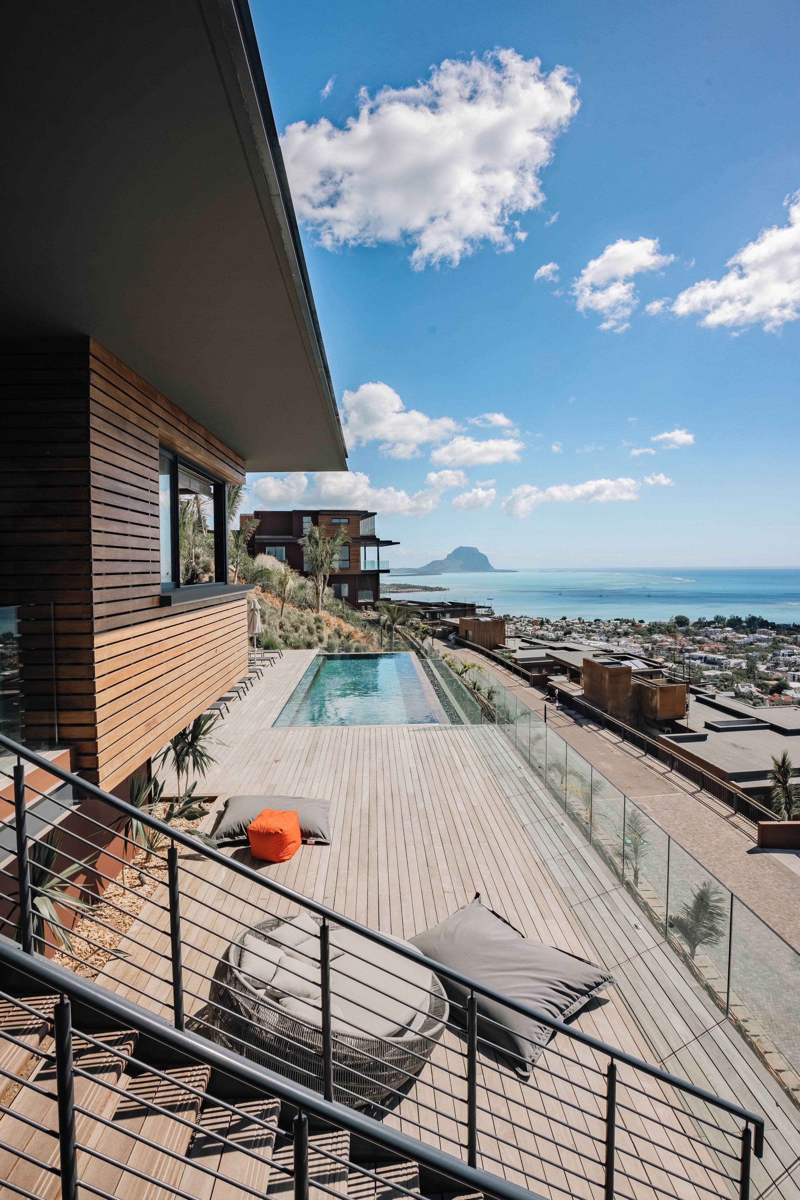A modern outdoor pool area with a wooden deck, lounge chairs, and a view of the sea and distant mountains.