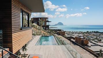 A modern outdoor pool area with a wooden deck, lounge chairs, and a view of the sea and distant mountains.